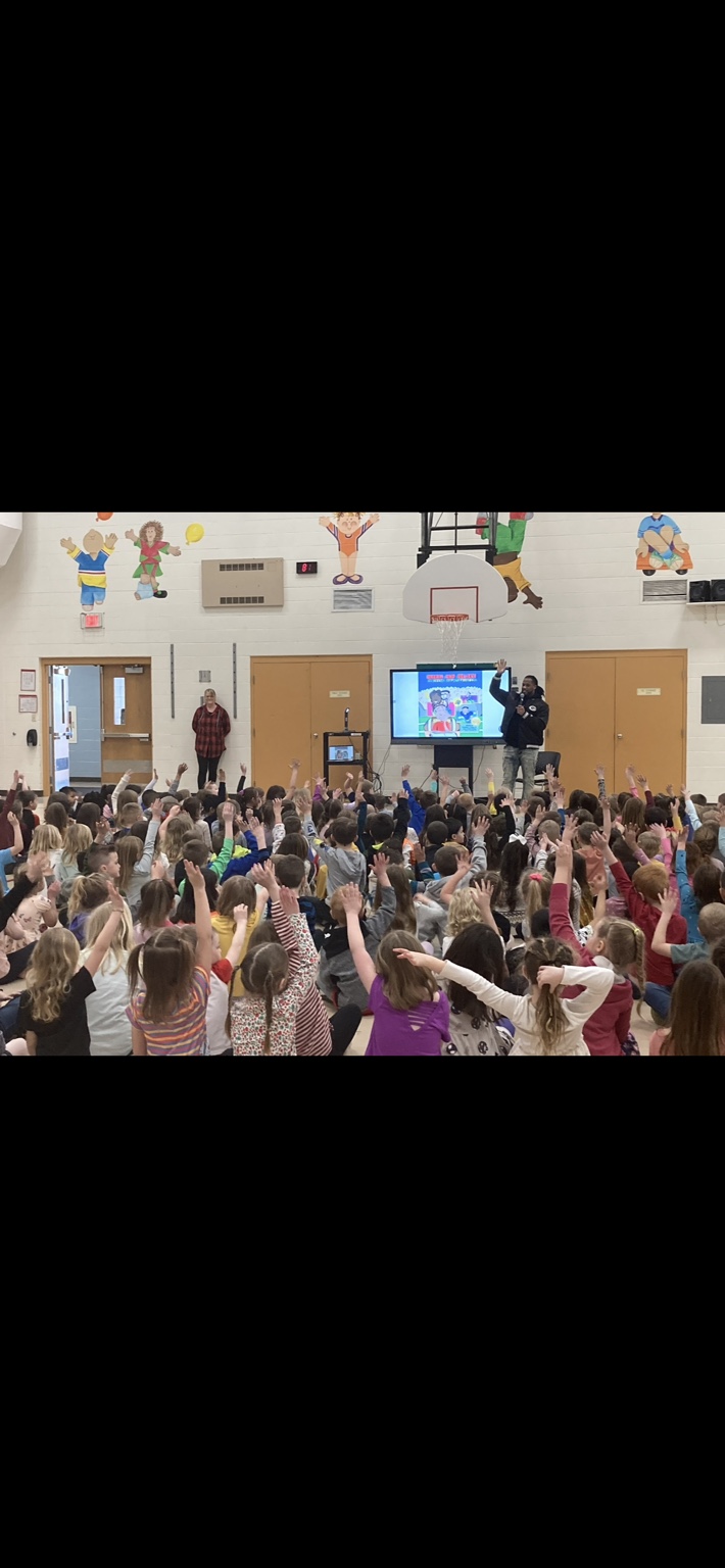Alex Green leading a school assembly with children raising their hands