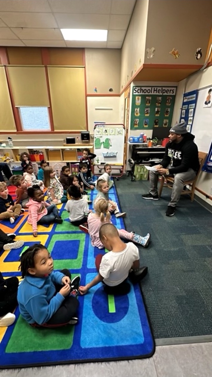 Alex Green reading to children seated on a colorful classroom rug